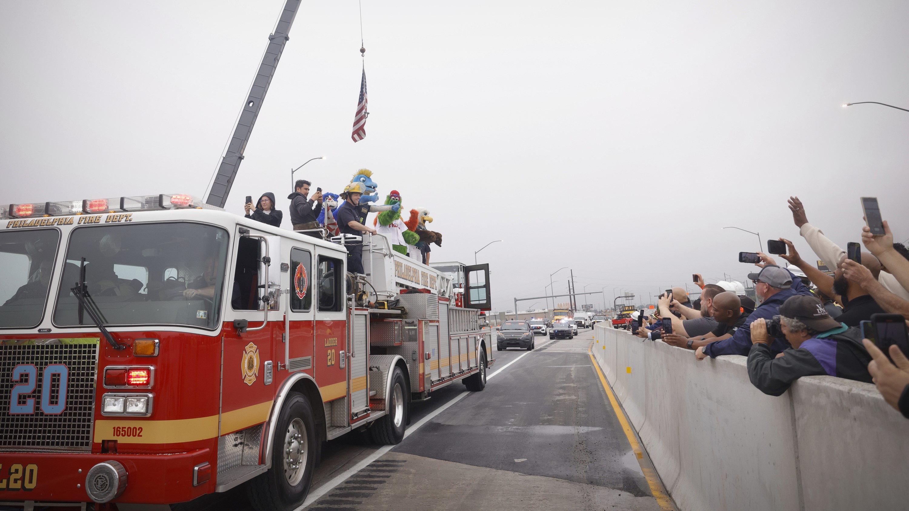 A fire truck and other vehicles drive over the temporary I-95 overpass on opening day while individuals standing behind concrete barriers take photos and wave; atop the fire truck stand firefighters as well as the Philly Philadelphia Phillies Phantic mascot and the Philadelphia Eagles mascot.