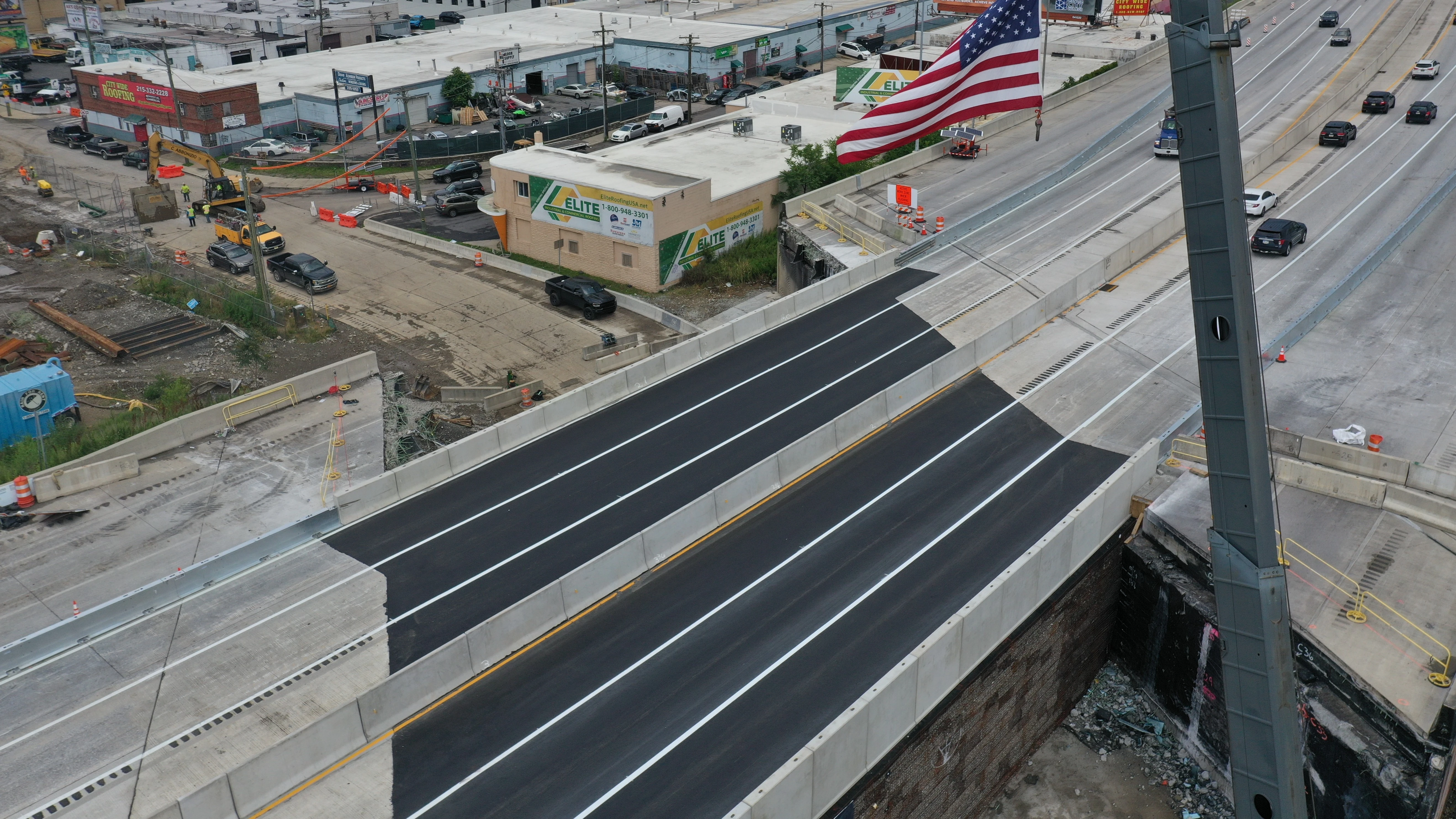 An aerial image of the temporary I-95 overpass structure on opening day with a United State flag affixed to a crane flying high above it.