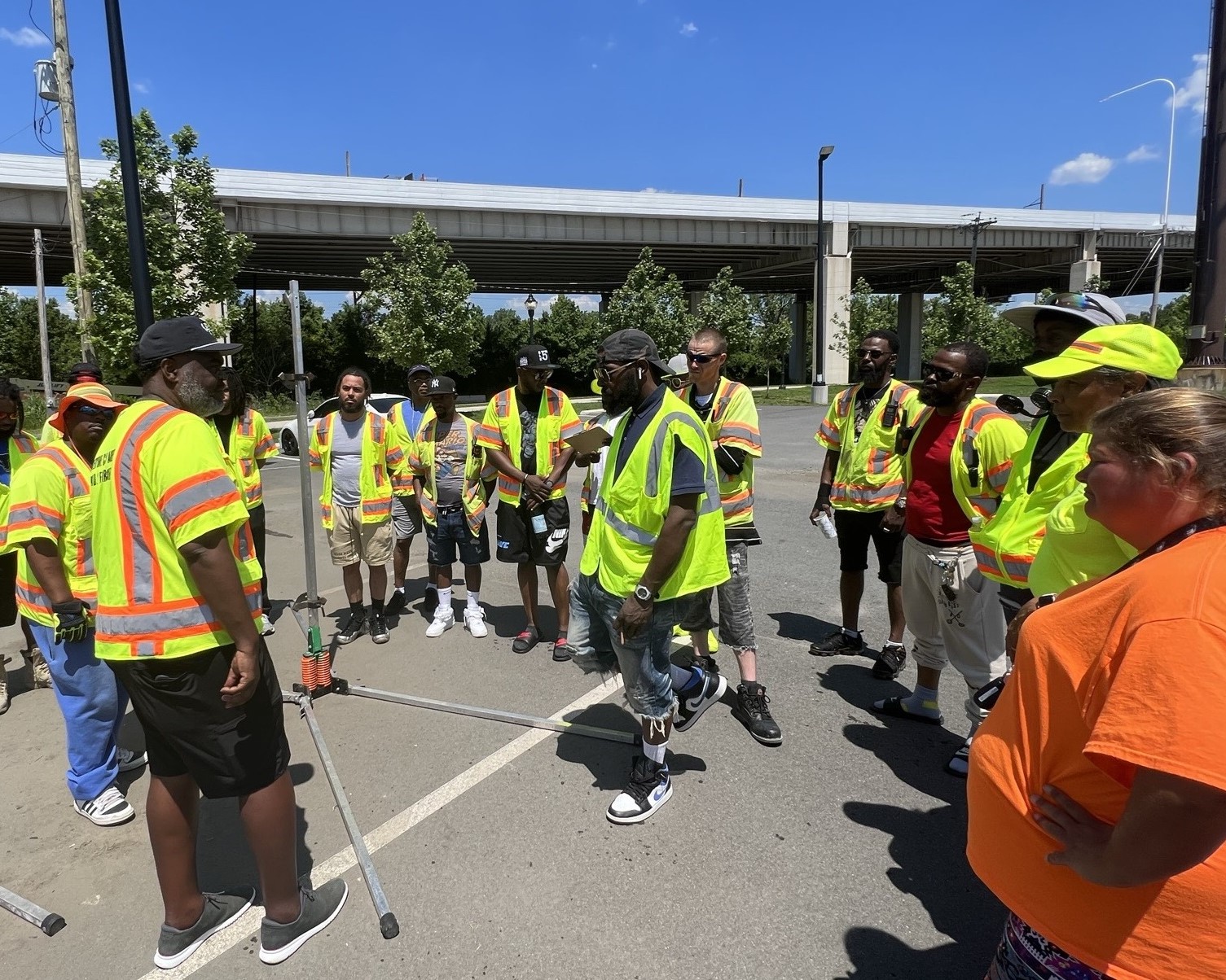 An image of a group of individuals in yellow safety vests standing in a parking lot next to a bridge, circled around an individual in a yellow safety vest who is talking to them