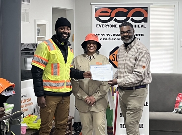 An image of three individuals in front of an Everyone Can Achieve (ECA) Corp orange, black and white banner, as one of the individual's hands one of the other individuals a certificate of achievement