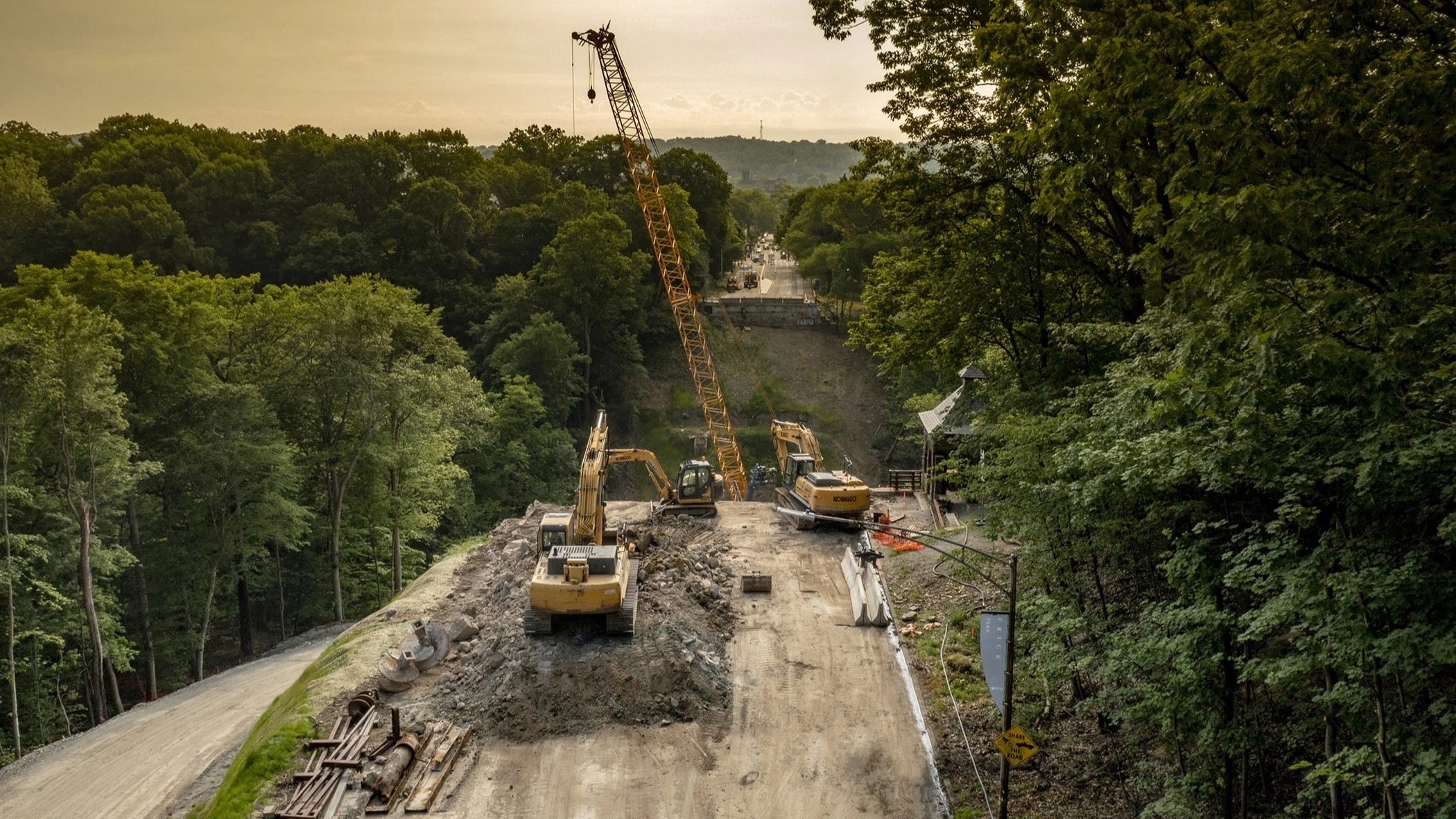 An image of large yellow pieces of construction equipment in the foreground next to an open space where the Fern Hollow Bridge used to prior to the collapse; the site is being prepared for the rebuilding of the bridge 