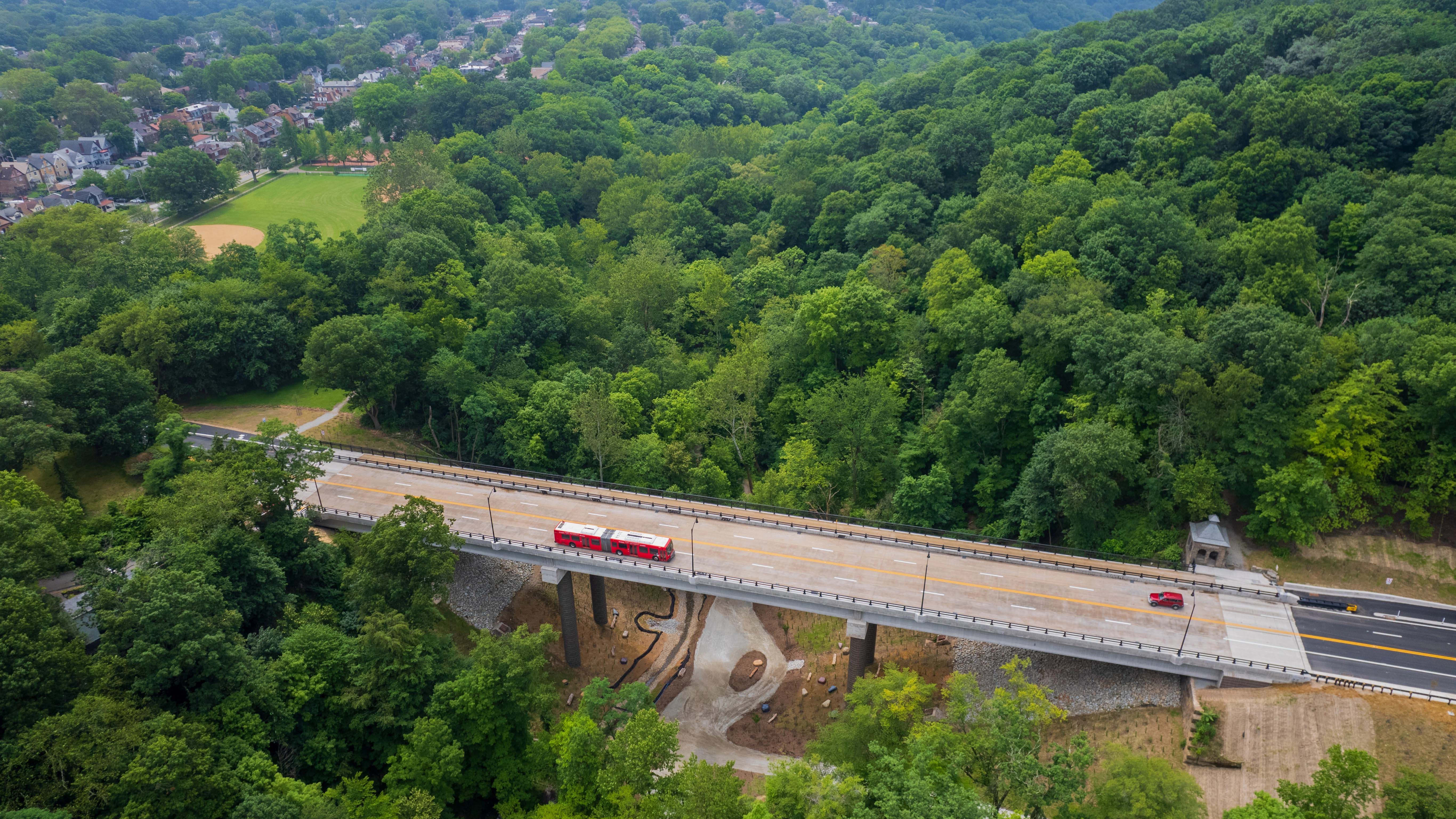 An aerial image of a red Pittsburgh Regional Transit bus and a red SUV crossing the completed Fern Hollow Bridge 