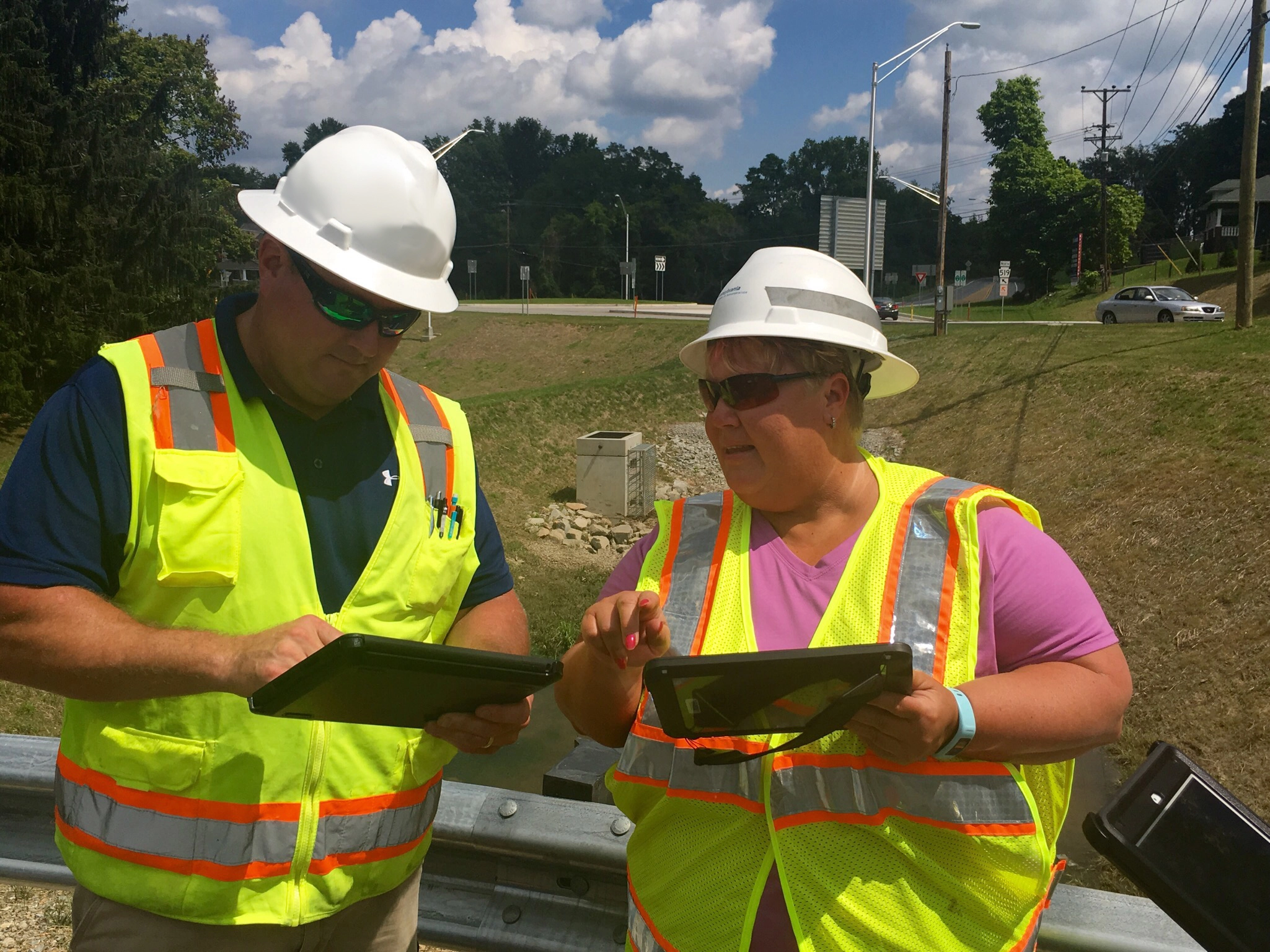 An image of two workers in white hard hats and yellow safety vests standing next to each other at a job site, talking and entering information about a construction projects onto their mobile devices