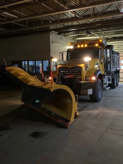 An image of a yellow PennDOT plow truck inside a garage facility with its lights on and fluorescent orange markers affixed to either side of the plow blade