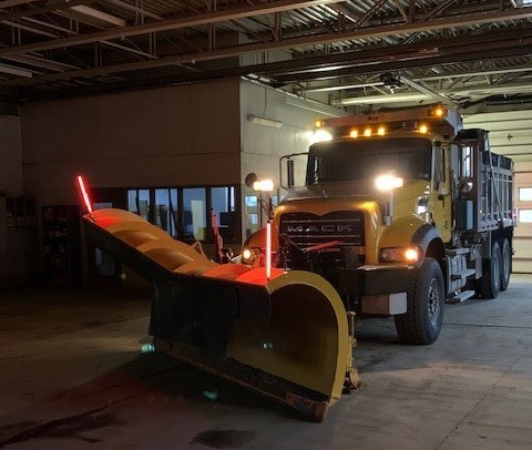 An image of a yellow PennDOT plow truck inside a garage facility with its lights on and fluorescent orange markers affixed to either side of the plow blade