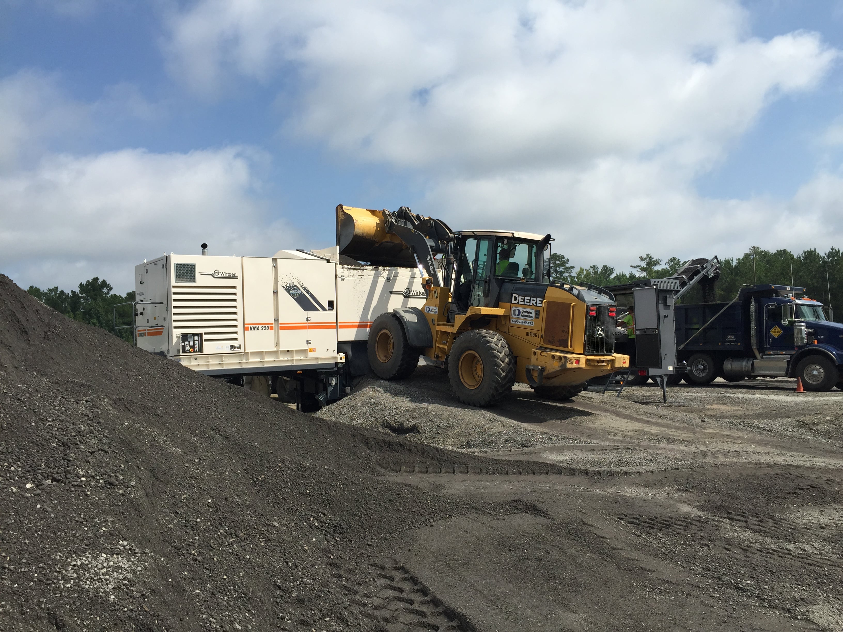 An image of a worker operating a backhoe to place Reclaimed Asphalt Pavement (RAP) in a white metal mobile cold recycling plant where it is mixed with a liquid, such as asphalt emulsion or foamed asphalt, and a small amount of cement to create a new flexible asphalt base