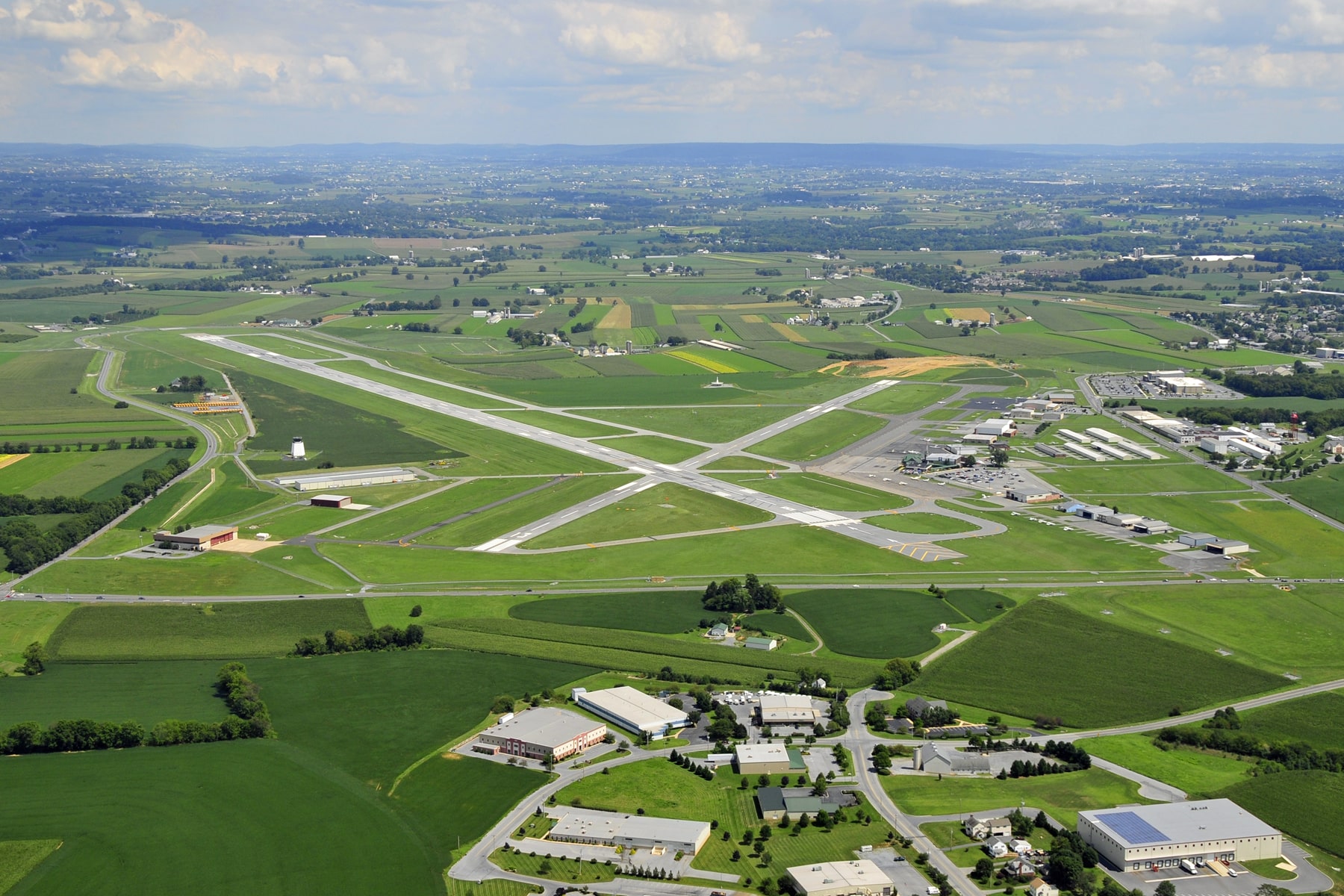 An aerial image of an airport runway with green fields, houses, and larger buildings visible in the surrounding area