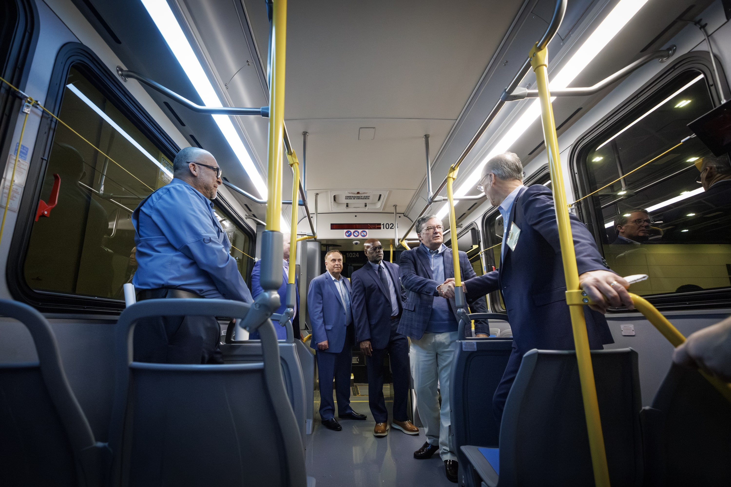 An image of five men in suits standing inside a public transit bus shaking hands and talking with one another