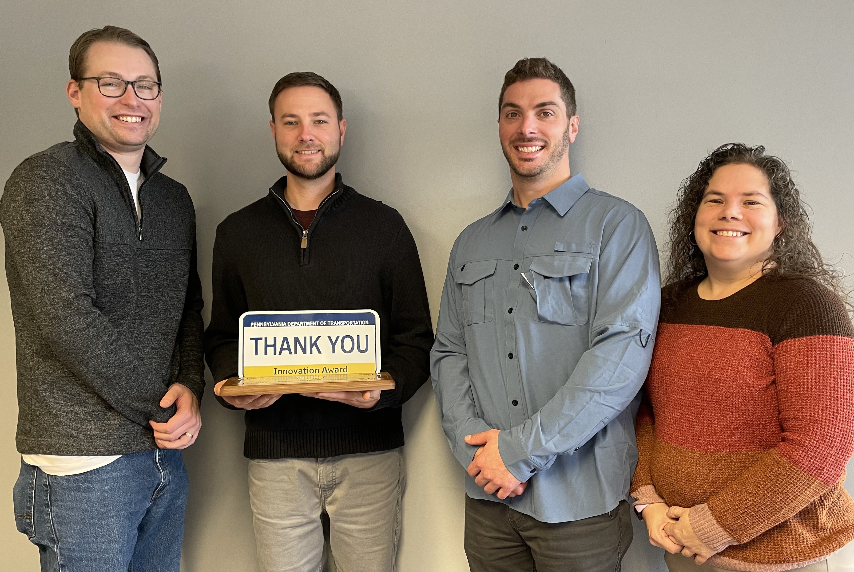 Four individuals stand in a line smiling with one of the individuals holding an award that looks like a blue, yellow, and white Pennsylvania license plate with the words 'Thank You' on it